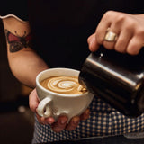 Person pouring coffee into a white cup with a tattooed arm and checkered sleeve.